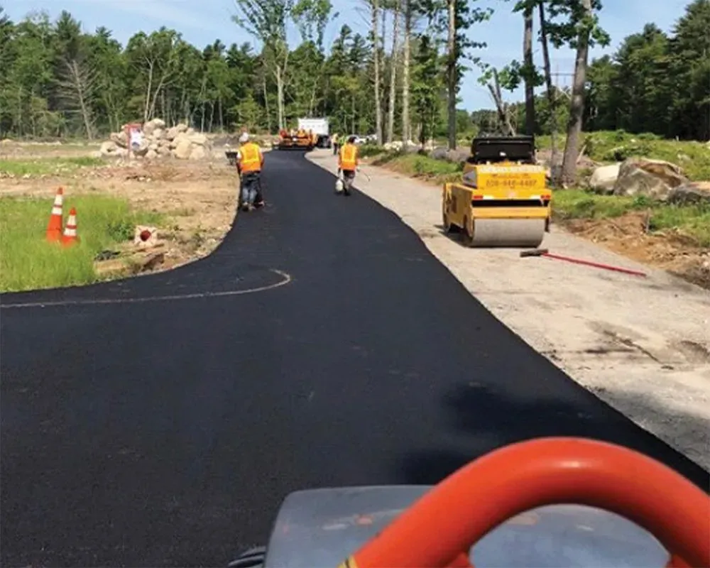 Workers paving a road. Two workers use hand rollers. A roller compactor and construction equipment are visible.