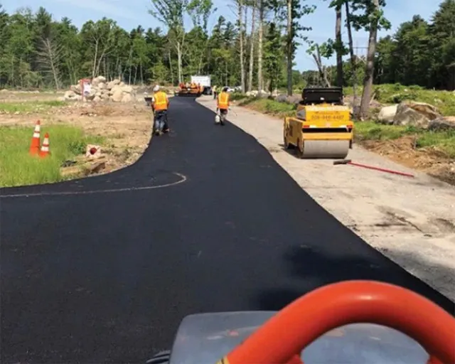 Workers paving a road. Two workers use hand rollers. A roller compactor and construction equipment are visible.