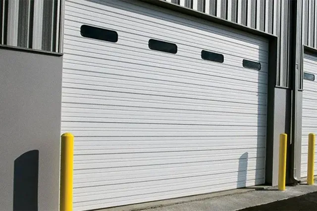 White industrial roll-up garage door with small windows, flanked by yellow safety bollards.
