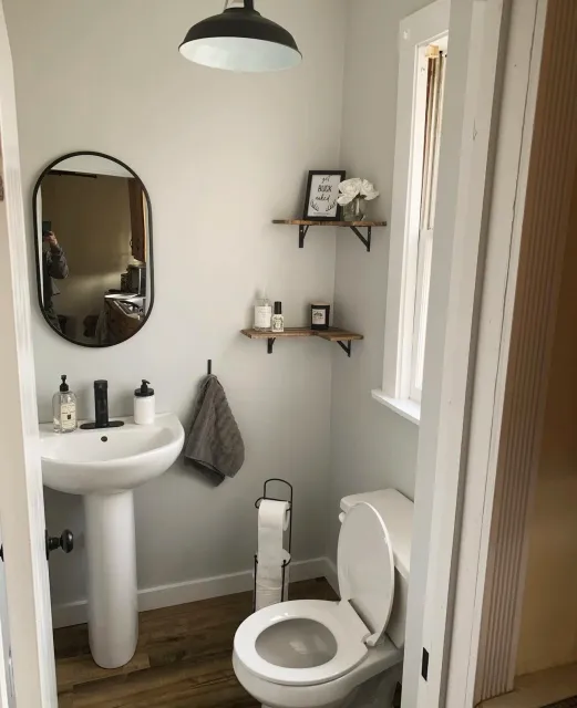 A small bathroom with a pedestal sink, oval mirror, black pendant light, and wooden shelves with decor. Toilet paper holder and window are present. Bathroom supplies on sink and shelves.