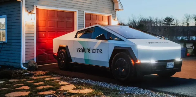 A futuristic white truck labeled venturehome. is parked in front of a red garage at dusk, with house lights illuminating the scene.