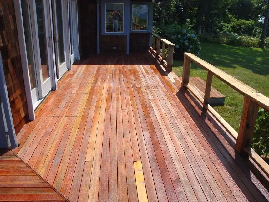 A long, wooden outdoor deck with reddish-brown planks, a railing, and a small step, attached to a house with glass doors.
