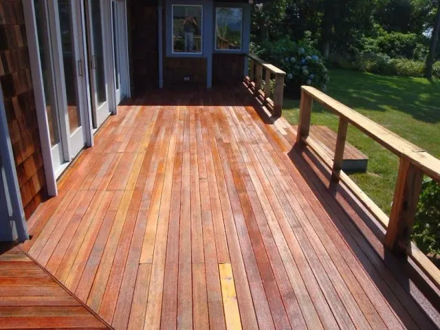 A long, wooden outdoor deck with reddish-brown planks, a railing, and a small step, attached to a house with glass doors.