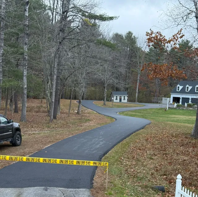 Newly paved black asphalt driveway curves through a wooded area. Yellow caution tape is across the foreground.