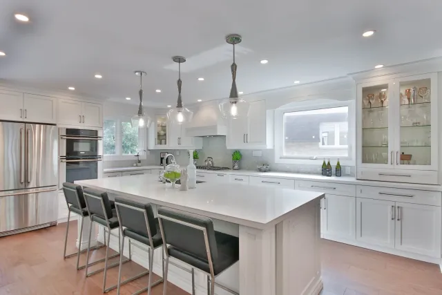 Modern white kitchen with large island, black chairs, stainless steel refrigerator, built-in oven, and pendant lighting, with natural light coming through windows.