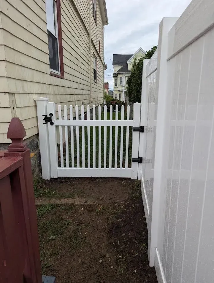 Narrow side yard with a white picket gate between a beige house and white fence.