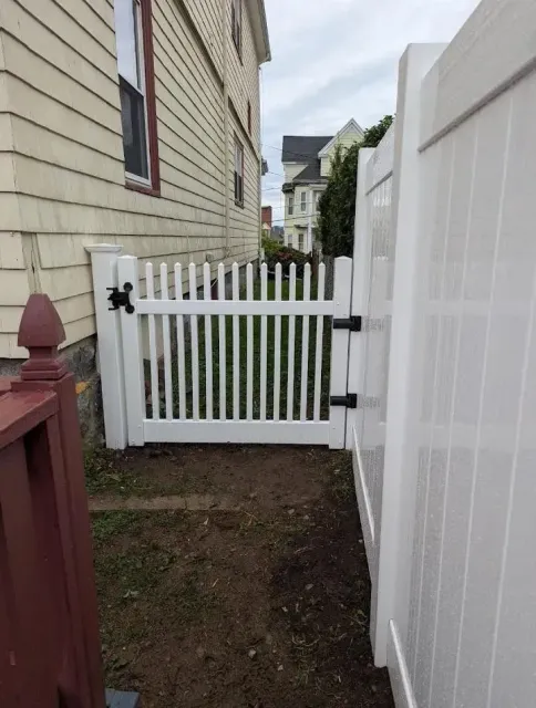Narrow side yard with a white picket gate between a beige house and white fence.