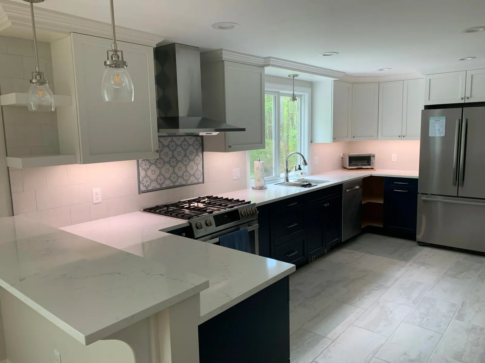 Modern kitchen with white countertops, black lower cabinets, light gray upper cabinets, stainless steel appliances, a window above the sink, and pendant lights.
