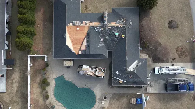 Aerial view of a house with damaged roof, debris in the yard, and a swimming pool nearby
