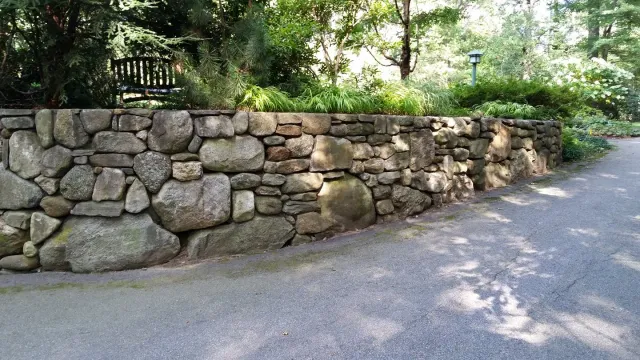 Stone wall bordering a gravel path, with greenery and trees in the background.