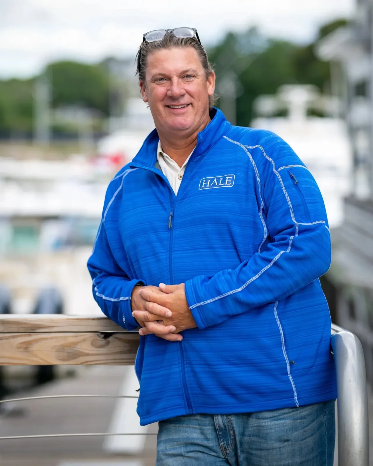 A man in a navy blue hoodie and a gray cap with 'HALE' written on it, standing outdoors near a wooden railing with a marina in the background.