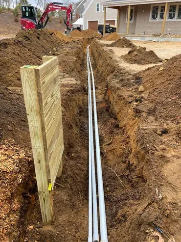 A construction trench with white pipes, a wooden support, and a backhoe in the background.