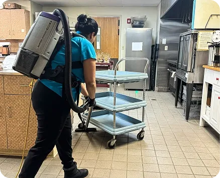 Commercial cleaning specialist using a backpack vacuum cleaner and pushing a utility cart in a large commercial kitchen facility.