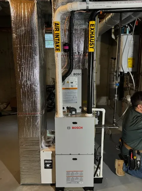 A Bosch furnace with air intake and exhaust pipes. A worker is in the frame. The setting appears to be a basement.