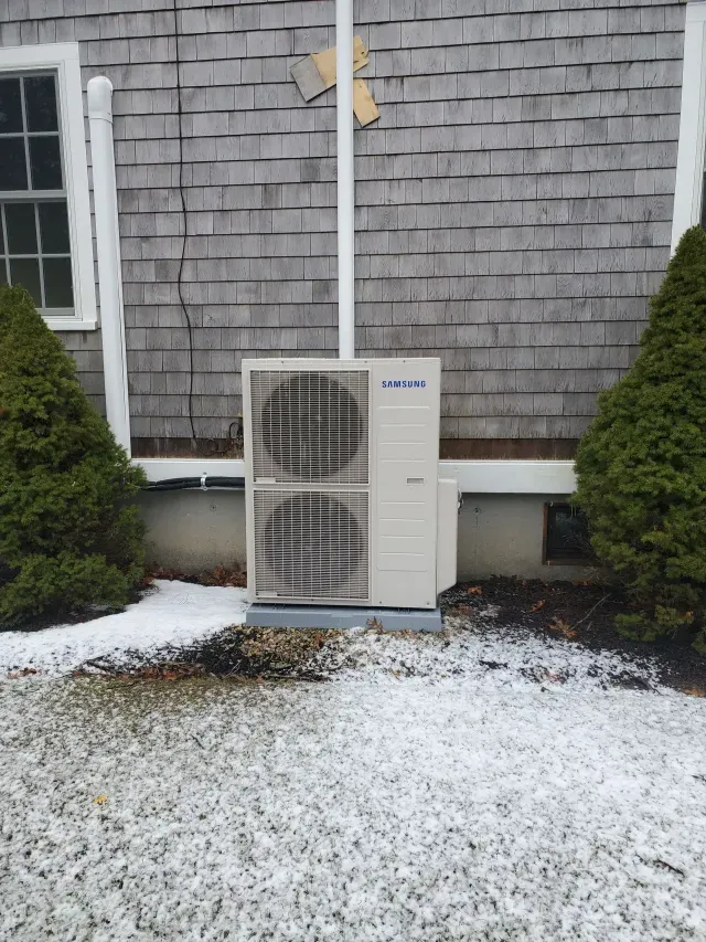Heat pump unit outside a house with cedar shingles, in a snowy setting.