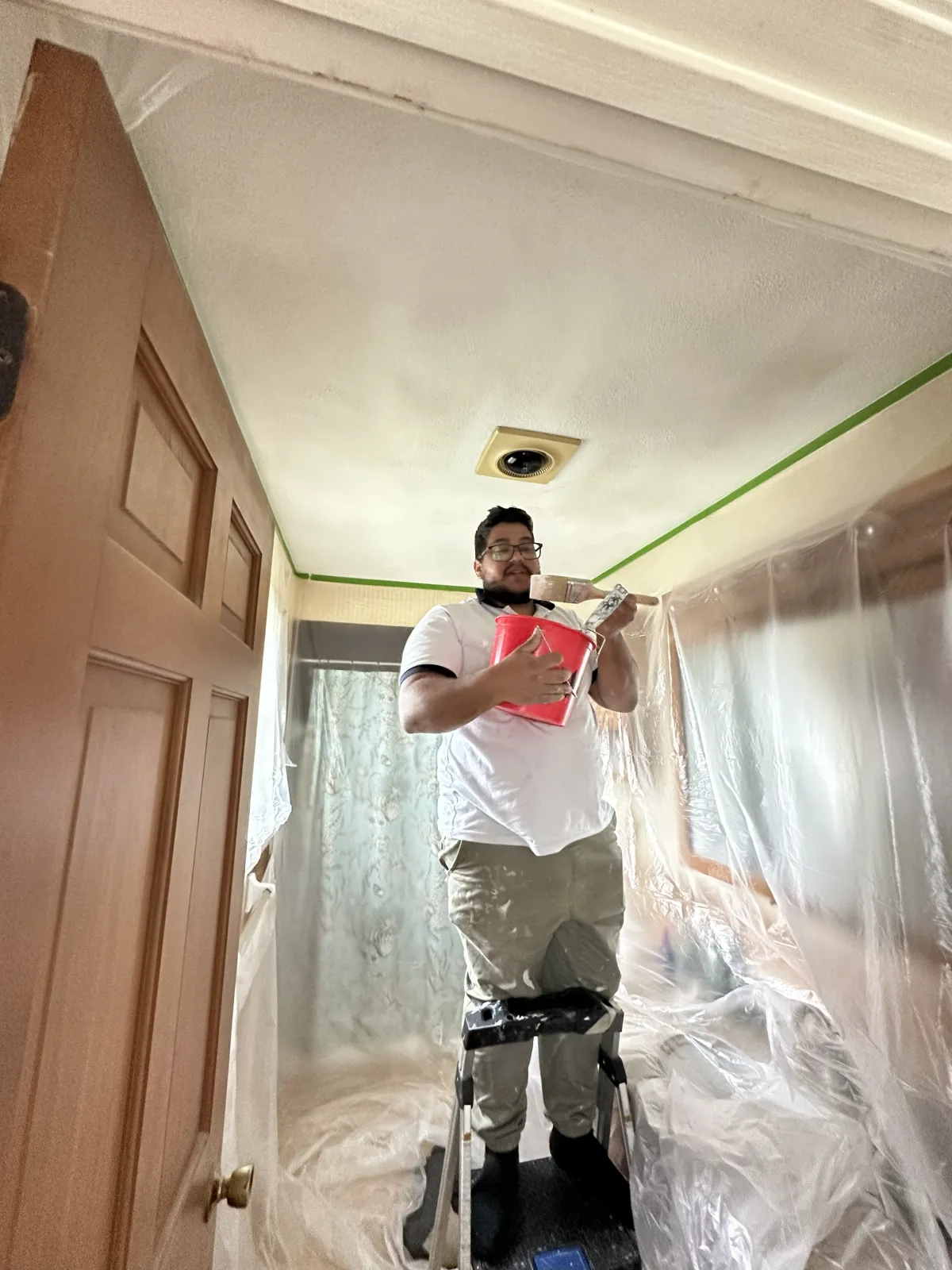A man standing on a step stool in a room, holding a red bucket and painting the ceiling while smiling. The room is covered with plastic sheets for painting.