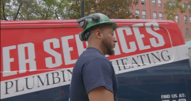 A man wearing a green hard hat walks past a red and blue van with "PLUMBING" and "HEATING" partially visible on the side.