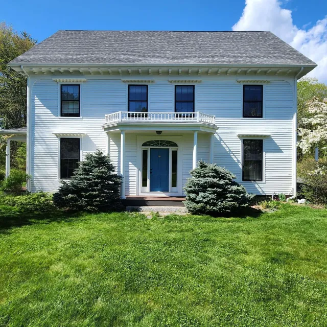White two-story colonial house with blue shutters, front porch, and a green lawn under a clear blue sky