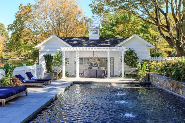 Backyard with a pool, white house with a porch, outdoor seating, and lush trees.