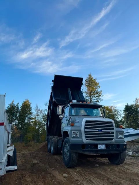A large dump truck is dumping dirt on a construction site with trees and a clear blue sky in the background.