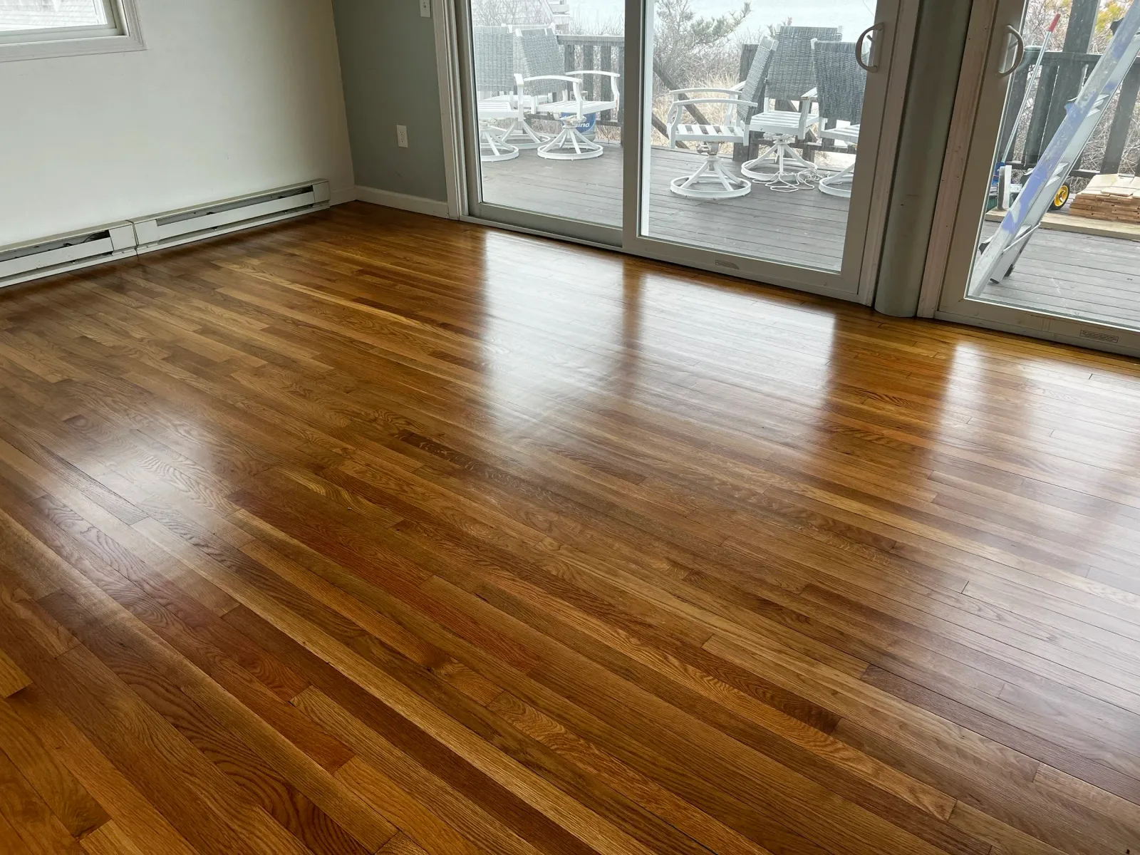 Room with polished hardwood floor, sliding glass doors leading to an outdoor deck with outdoor furniture, and a ladder outside.
