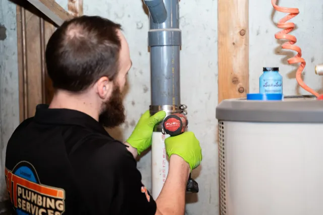 A man wearing a black shirt with a plumbing service logo and bright green gloves is working on a plumbing pipe with a cordless drill in a basement or utility area.