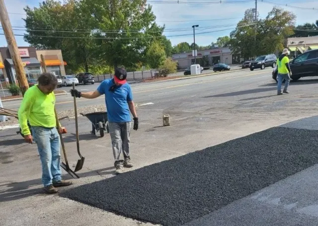Workers paving a street, spreading fresh black asphalt near a wheelbarrow on a sunny day