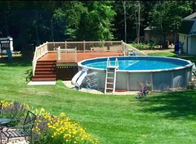 Above ground pool with a wooden deck, surrounded by green grass and trees in a backyard setting.