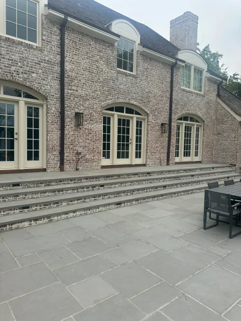 Back of a brick house with patio steps leading up to large glass doors and windows, outdoor dining table with chairs, and a cloudy sky.