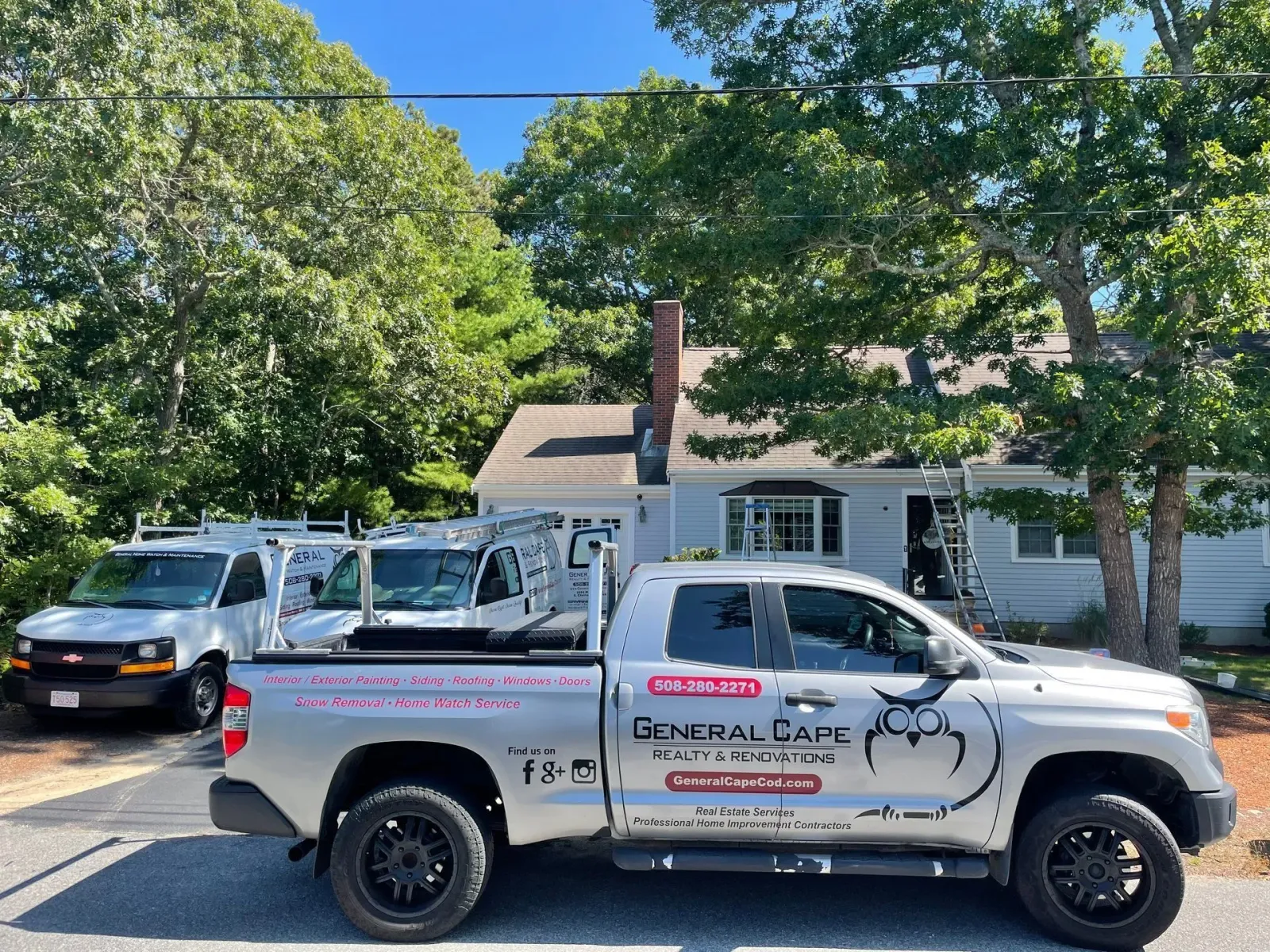 Three work vehicles parked in front of a light blue house with a chimney and trees, on a sunny day.