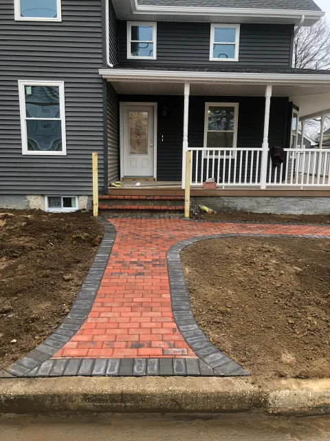 A new red brick walkway with a dark border leads to the front porch steps of a grey-sided house.