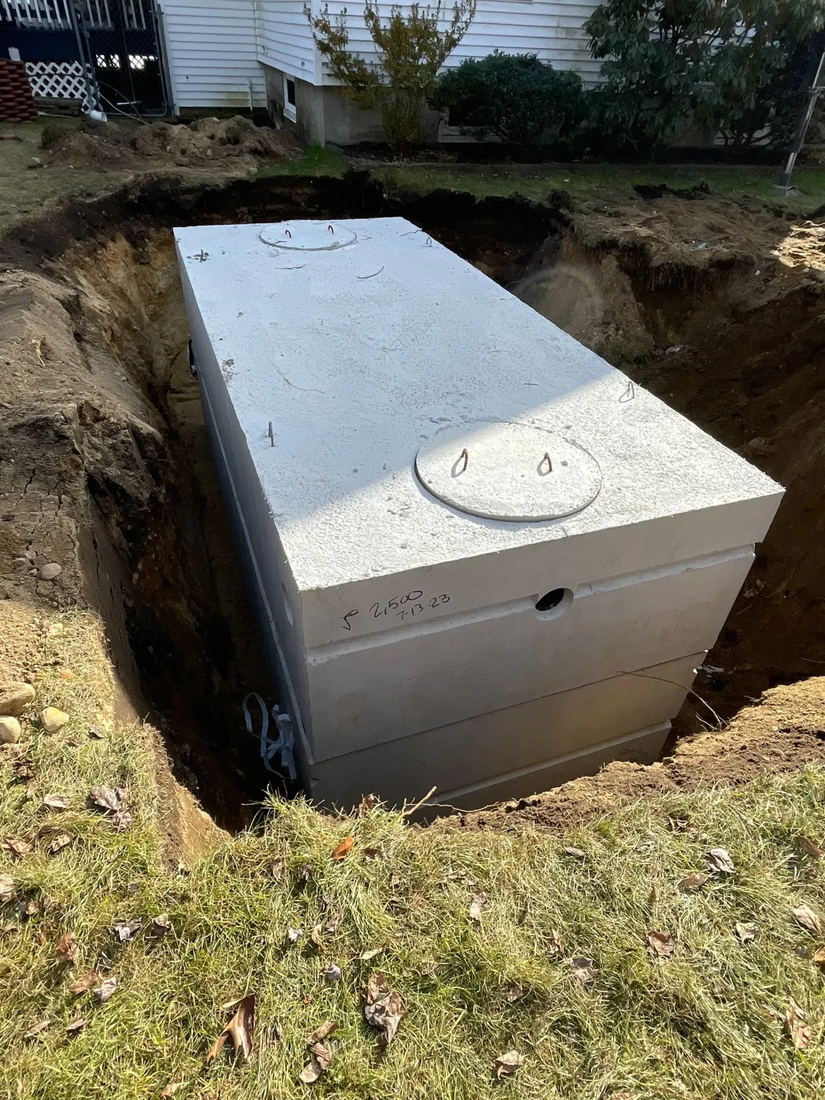 Large gray utility tank in a dug-out hole beside a house, with a round lid on top.