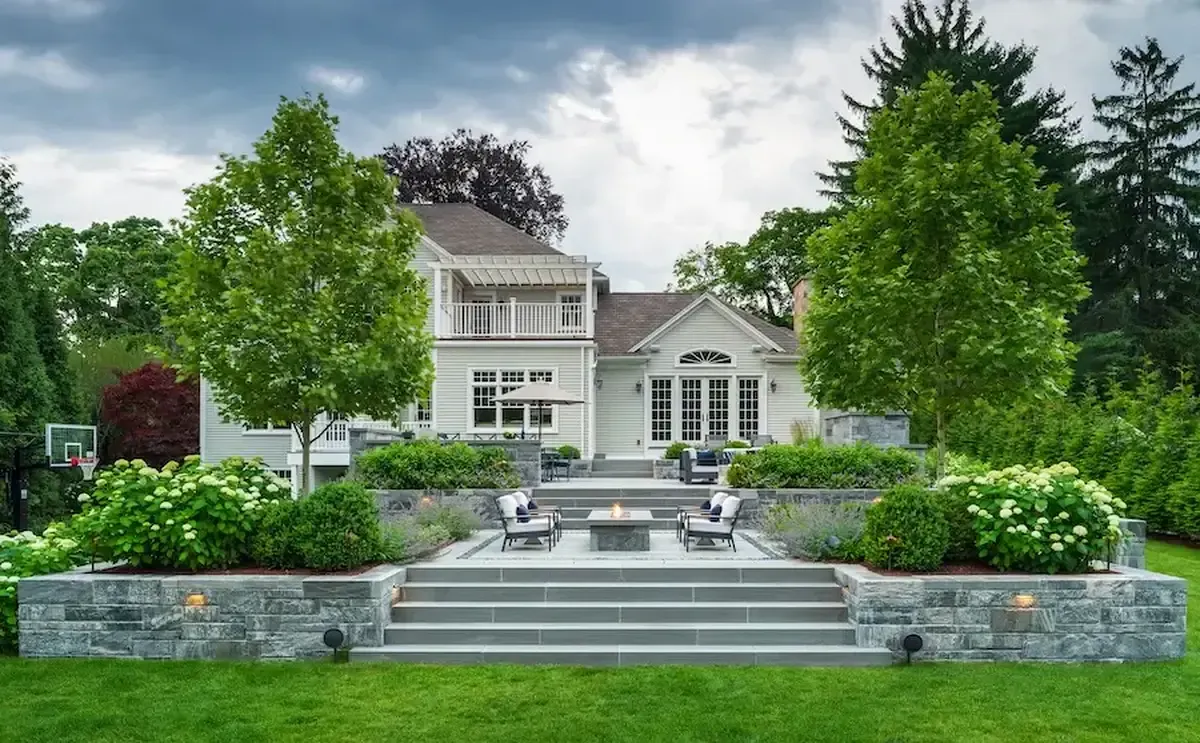 Backyard residence with a multi-tier stone patio, fire pit, and outdoor seating in front of a light-colored house surrounded by trees and shrubs.