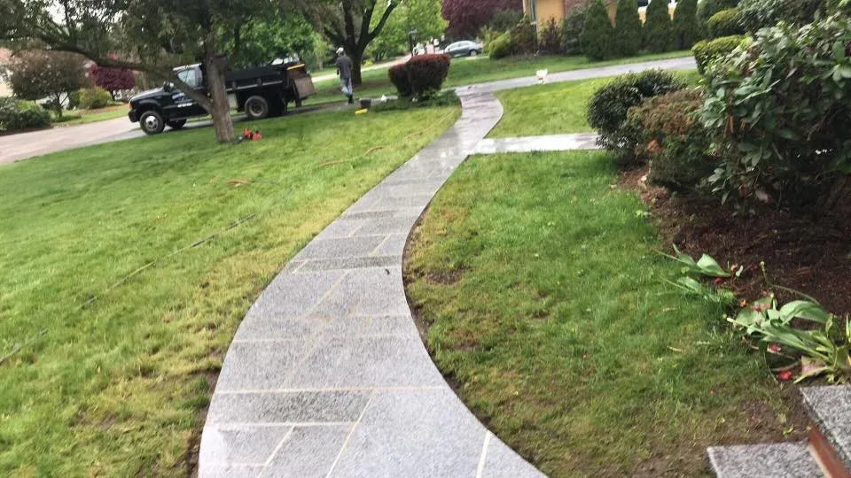 Curved concrete sidewalk running through a grassy lawn with shrubs and trees, and a parked truck in the background.