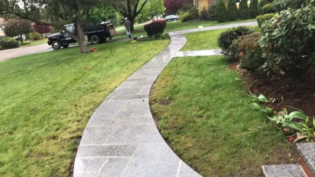 Curved concrete sidewalk running through a grassy lawn with shrubs and trees, and a parked truck in the background.