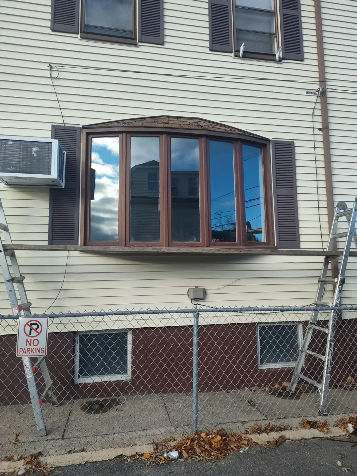 Damaged apartment exterior with broken bay window, bent siding, and chain-link fence in front