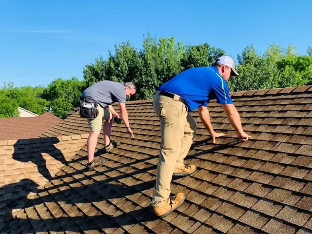 Two workers in safety gear inspect and work on a residential asphalt shingle roof on a sunny day