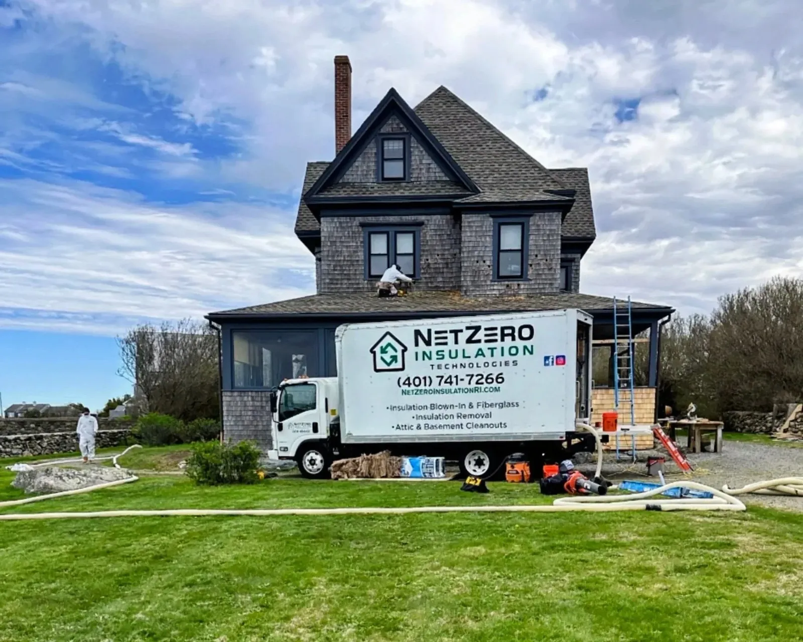 A house under repair with a sign for NetZero Insulation Technologies on a truck in front. Workers are working on the roof, and construction tools and materials are scattered on the yard.