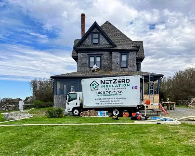 A house under repair with a sign for NetZero Insulation Technologies on a truck in front. Workers are working on the roof, and construction tools and materials are scattered on the yard.
