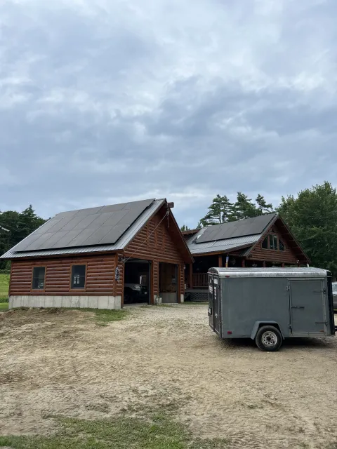 Log cabin with sloped roofs and solar panels, trailer parked in front, overcast sky.