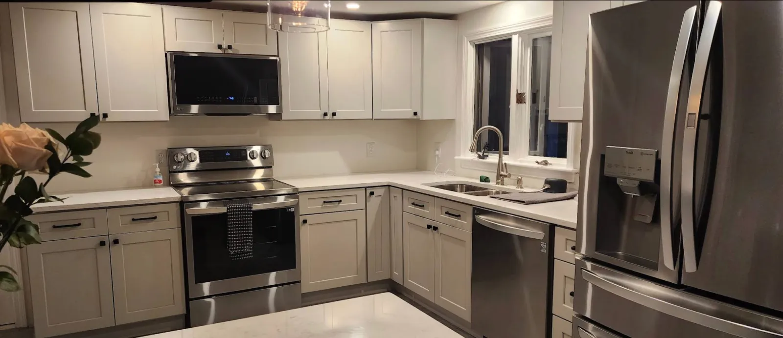 Modern kitchen with white cabinets, stainless steel appliances including a microwave, oven, and refrigerator, and a double sink near a window.