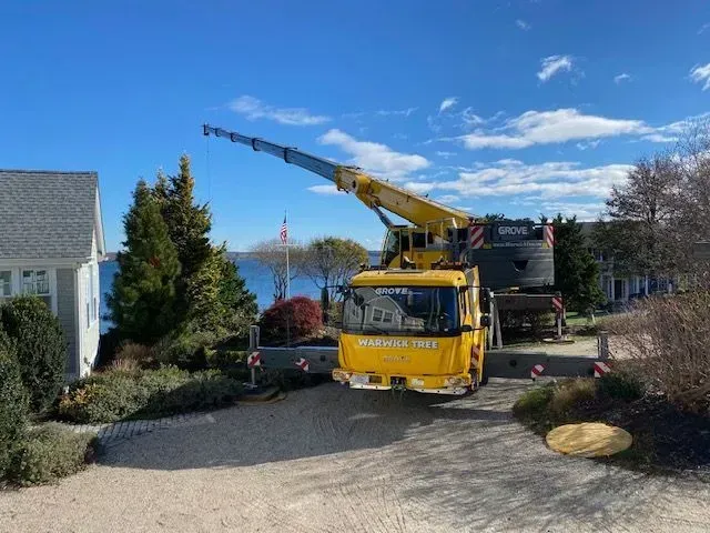 Yellow crane parked near a waterfront house, boom extended toward the water under a blue sky.
