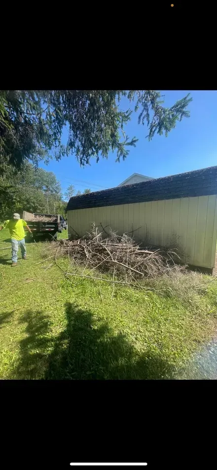 A person in a high-visibility vest clears a large pile of brush next to a long, light-colored fence on a sunny day.