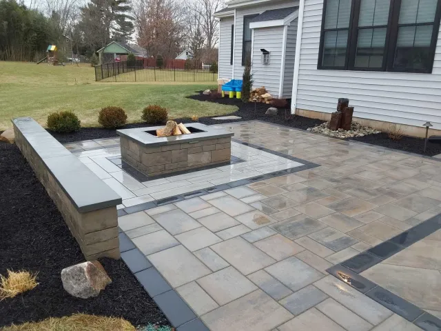 A backyard patio area with beige and gray stone tiles, a fire pit with wood fire inside, decorative rocks, a small water feature, and landscaping with shrubs and mulch next to a house with siding and large windows.