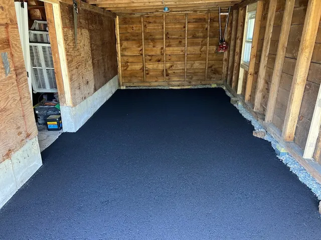 Inside of a garage; newly paved dark gray asphalt floor. Wooden walls and support beams.