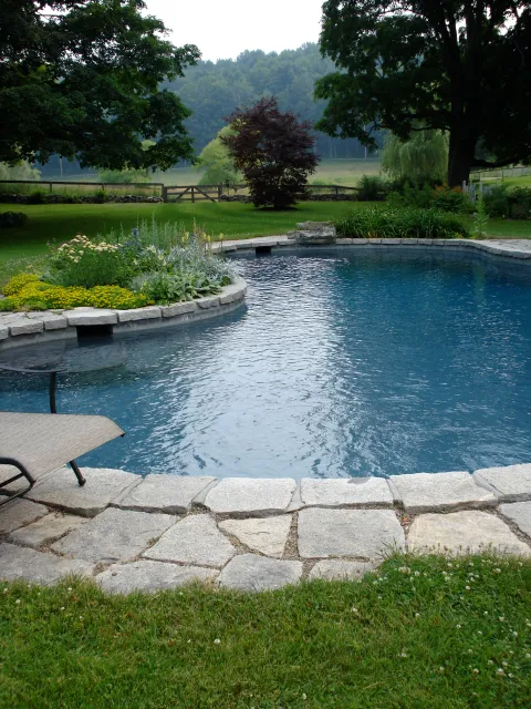 A backyard with a stone-paved swimming pool, surrounded by green grass and plants, with trees and rolling hills in the background.
