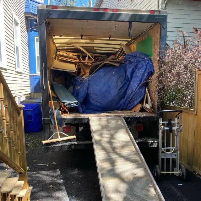 Truck bed loaded with debris, ramp extended. Blue tarp covers some items. Outdoors, next to a house.