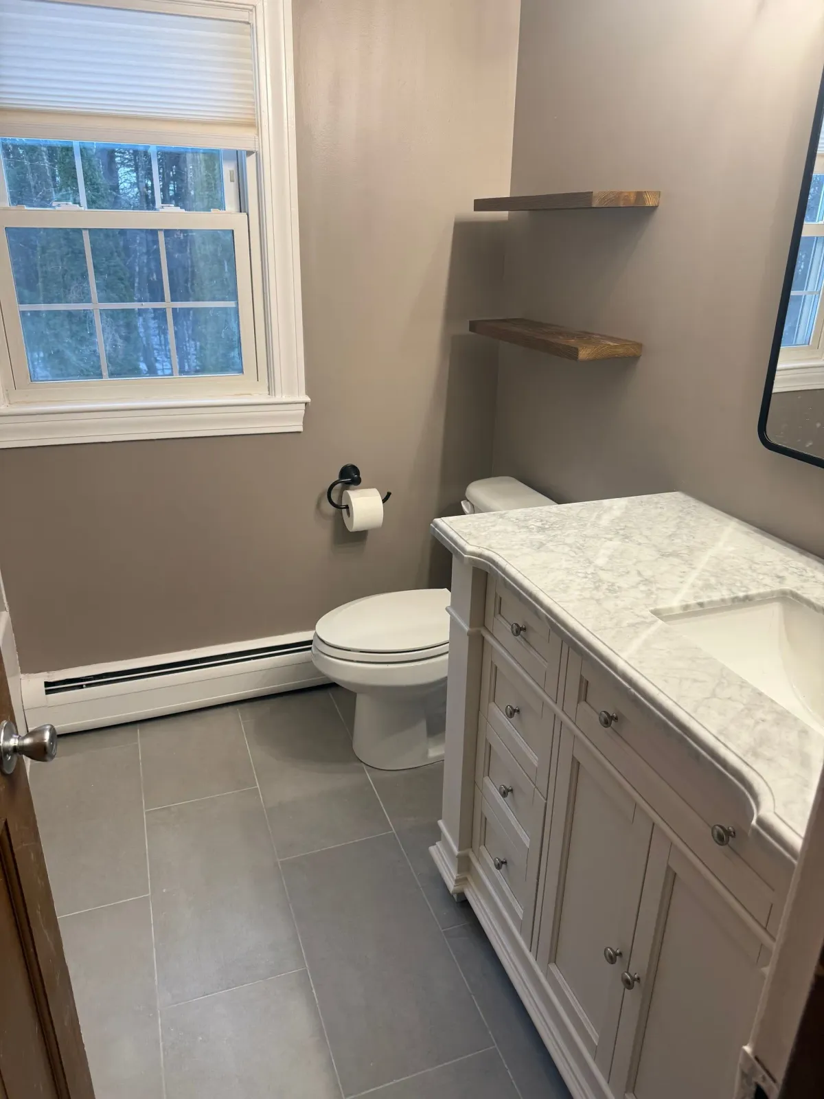 A bathroom with a toilet , sink , mirror and window.
