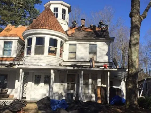 Old white Victorian house with a turret, damaged roof, and porch under a blue sky