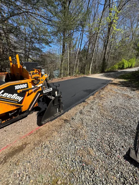 Orange skid-steer paving a black gravel path beside trees and gravel driveway